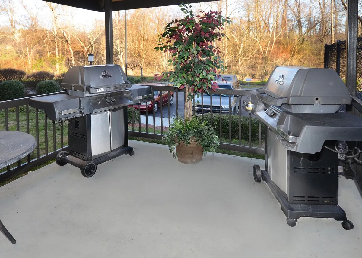 The scene showcases two outdoor grills in a well-landscaped, green setting, ideal for family barbecues or casual outdoor dining. The grills are surrounded by decorative plants and an open area that encourages socializing and enjoying the outdoors.