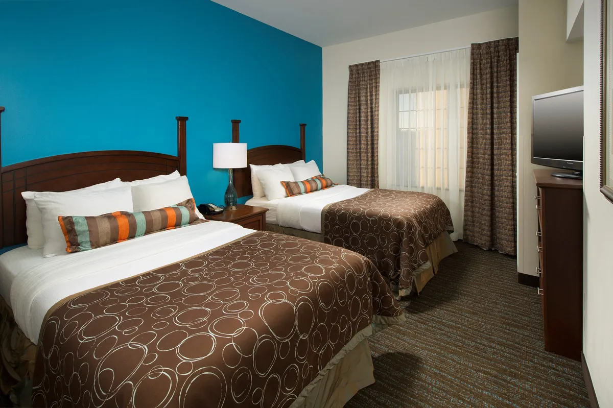 This image showcases a stylish suite with two double beds adorned with patterned blankets, a vibrant blue accent wall, a modern bedside lamp, curtains letting in natural light, and a wall-mounted television. The composition highlights the room's comfortable, welcoming atmosphere and capability for both relaxation and entertainment.