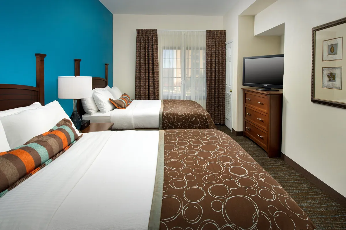 A bright and cheerful bedroom scene with two beds adorned with decorative pillows, a striking blue accent wall, a window dressed in sheer curtains allowing natural light, and wooden furnishings. The setup invites shared moments and relaxation, suited for various groups staying together.