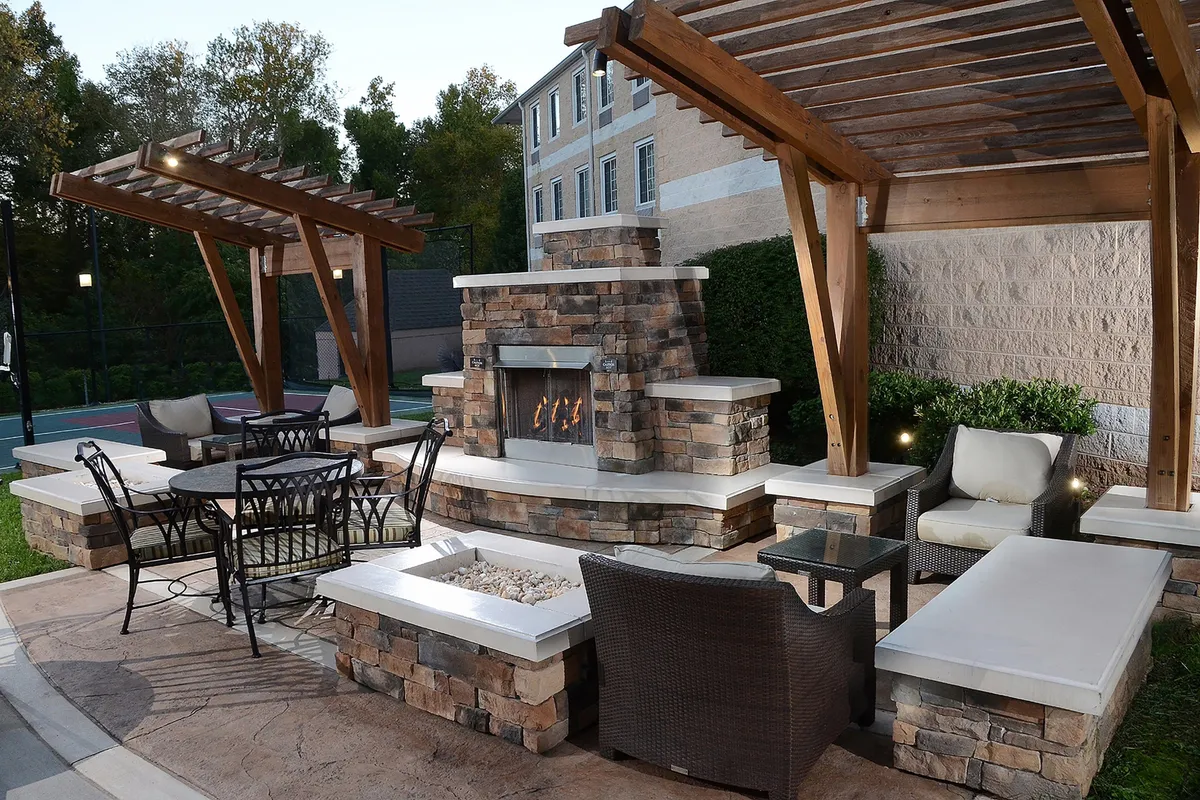 A warm, welcoming outdoor area featuring a glowing fireplace, comfortable seating, a pergola, and an outdoor dining table. Soft lighting and relaxed arrangements create a perfect setting for informal gatherings or solo unwinding after a busy workday.