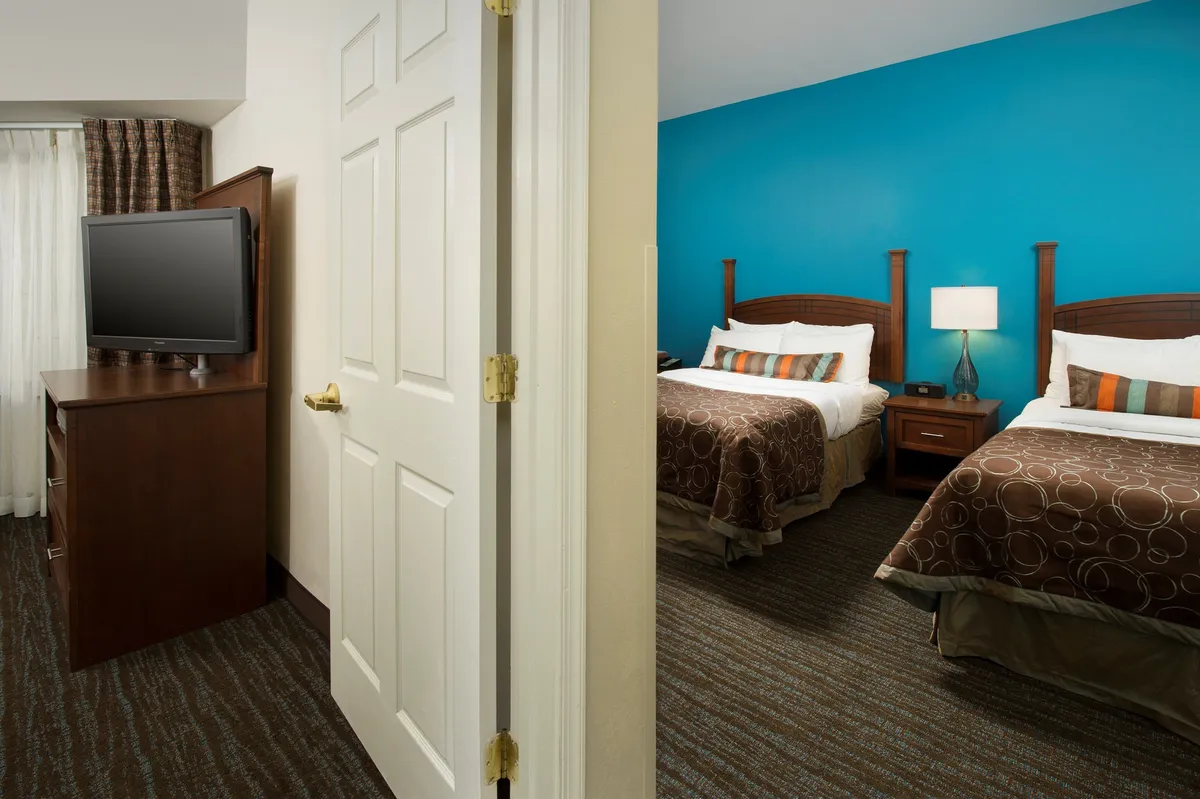 A welcoming bedroom interior featuring two double beds with crisp linens, a television set opposite the beds, and a colorful accent wall adding vibrancy to the space. The room exudes an inviting, restful atmosphere, perfect for winding down after a busy day. Blackout curtains can be noted, ensuring a dark and peaceful sleep environment.