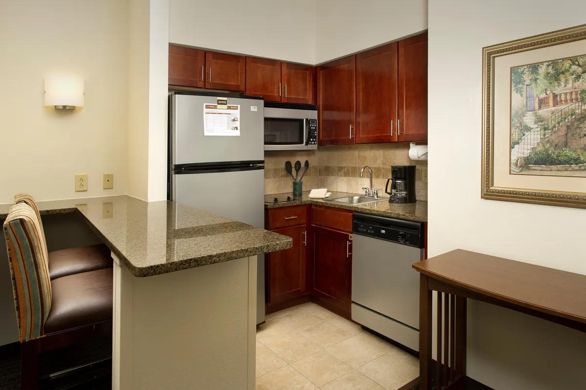 A cozy, functional in-suite kitchen featuring a kitchen bar, stainless steel appliances, wooden cabinetry, and decorative artwork. The space is filled with natural light and is designed for cooking, entertaining, and sharing meals.