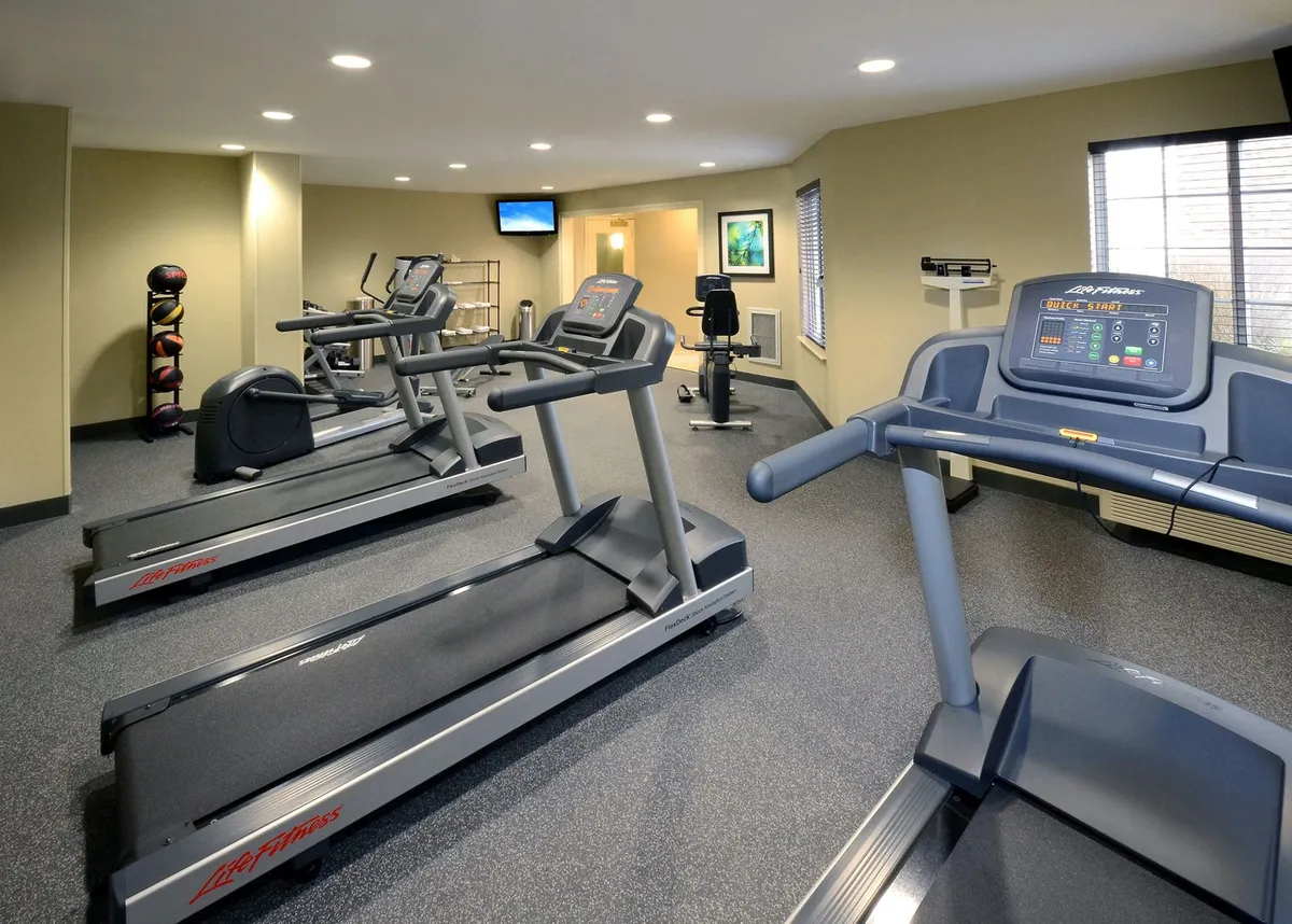 A modern fitness center featuring several treadmills, an elliptical machine, exercise balls, and a wall-mounted TV. The space feels bright and energizing, with ample room for guests to work out and socialize.