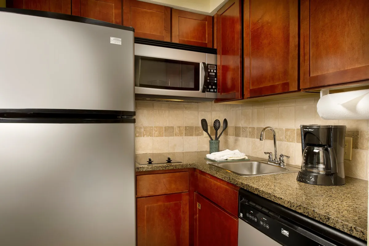 A close-up view of modern kitchen appliances including a stainless steel refrigerator, microwave, dishwasher, coffee maker, and kitchen sink. The setting is warm and inviting, reinforcing the practical amenities available for meal prep, coffee brewing, and snacking.