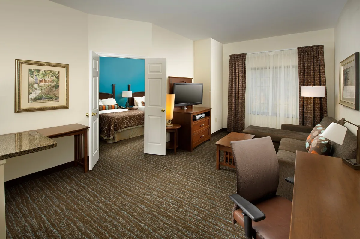 The image showcases a bright, inviting living area featuring a plush sofa facing a large TV, a convenient work desk, and an open view into the cozy bedroom. The scene exudes comfort and functionality, offering spaces to relax, work, and unwind all within arm’s reach.