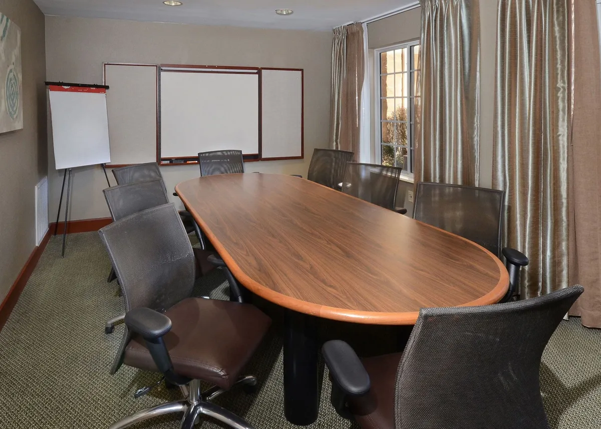 An interior business center with a long wooden conference table, ergonomic chairs, a whiteboard, and a large window flooding the room with natural light. The setup is ideal for meetings, collaborative work, or catching up on business tasks.