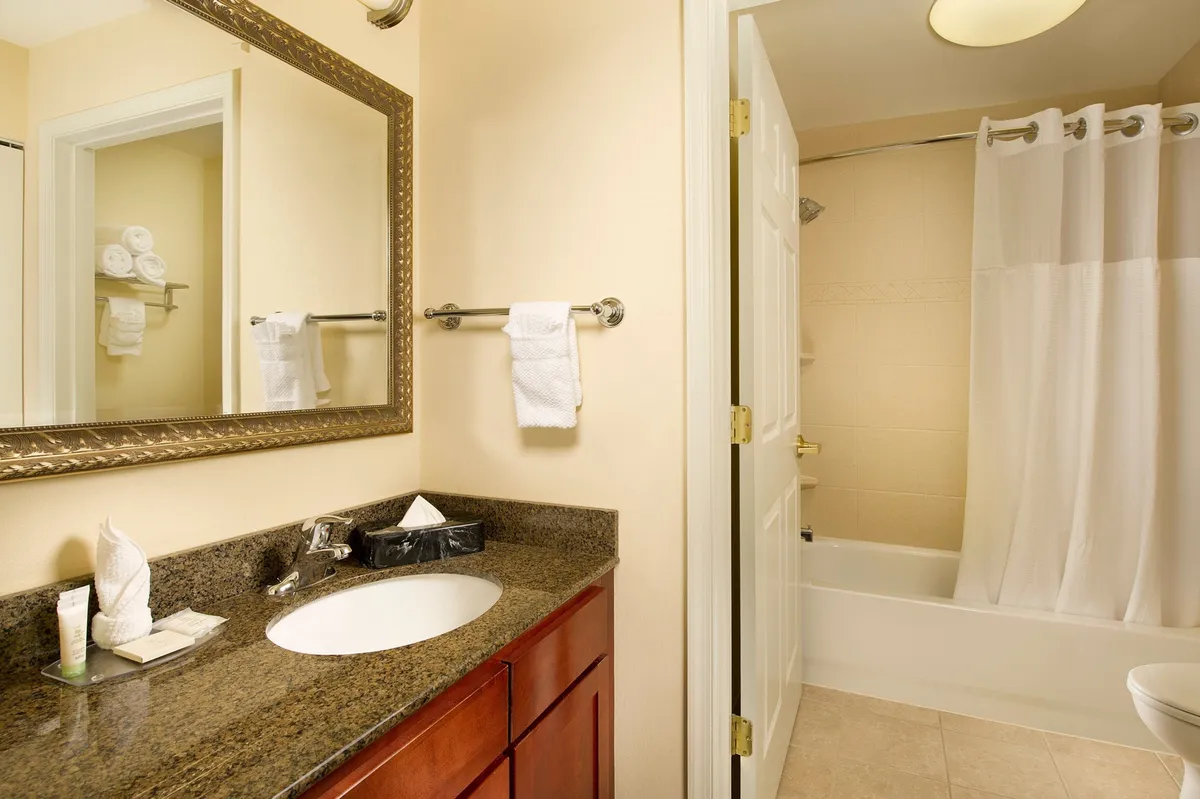 A modern bathroom interior featuring a granite countertop, large mirror, visible shower area, and neatly arranged towels on a rack. The space is bright, clean, and visually communicates comfort and easy self-care, aligning with the amenities mentioned in the text.