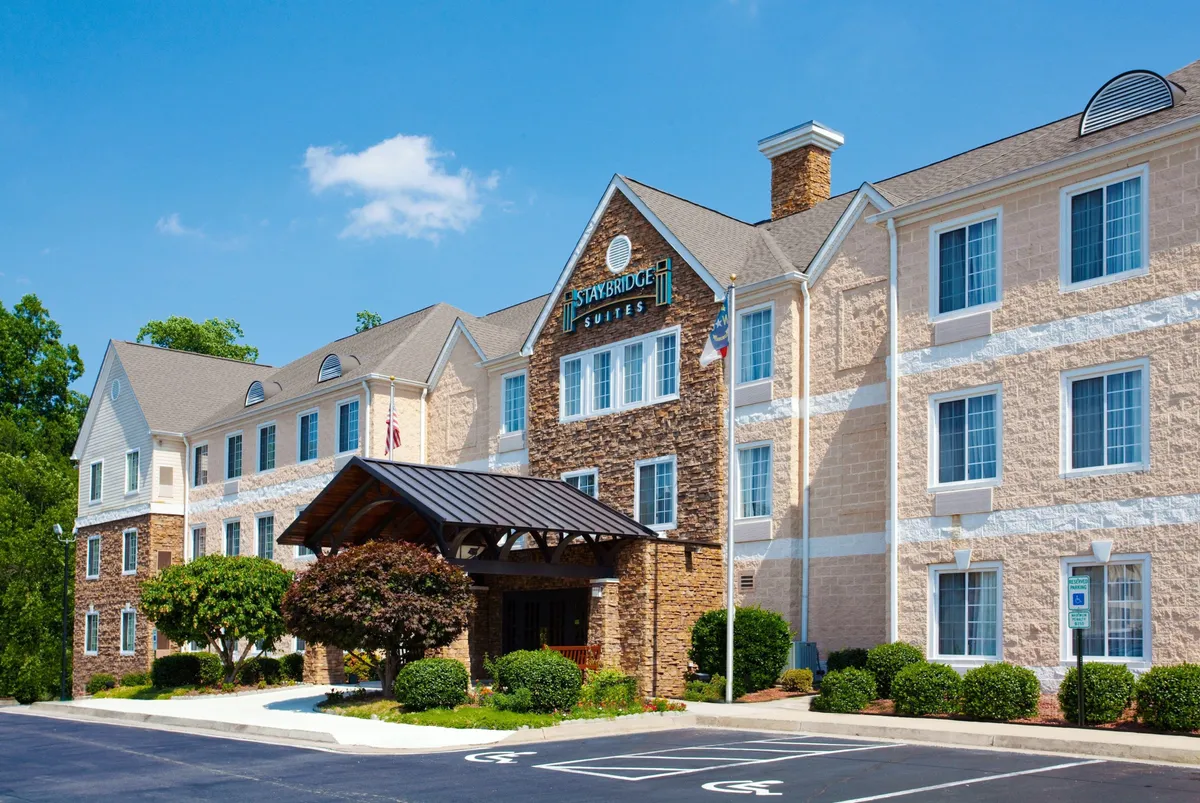 The image displays the inviting exterior of the Staybridge Raleigh Arpt-Morrisville, showing the entrance awning, stone facade, lush landscaped greenery, and prominent hotel signage. Guests can be seen arriving and relaxing near the entrance, setting a welcoming tone.